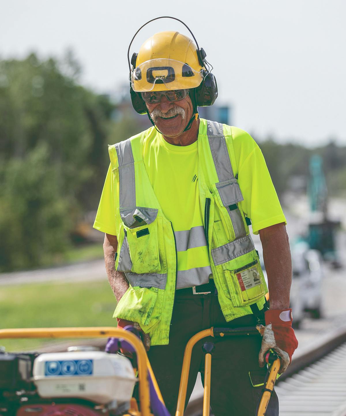 Smiling man, wearing safety glasses, hearing protection and hardhat, moving heavy equipment
